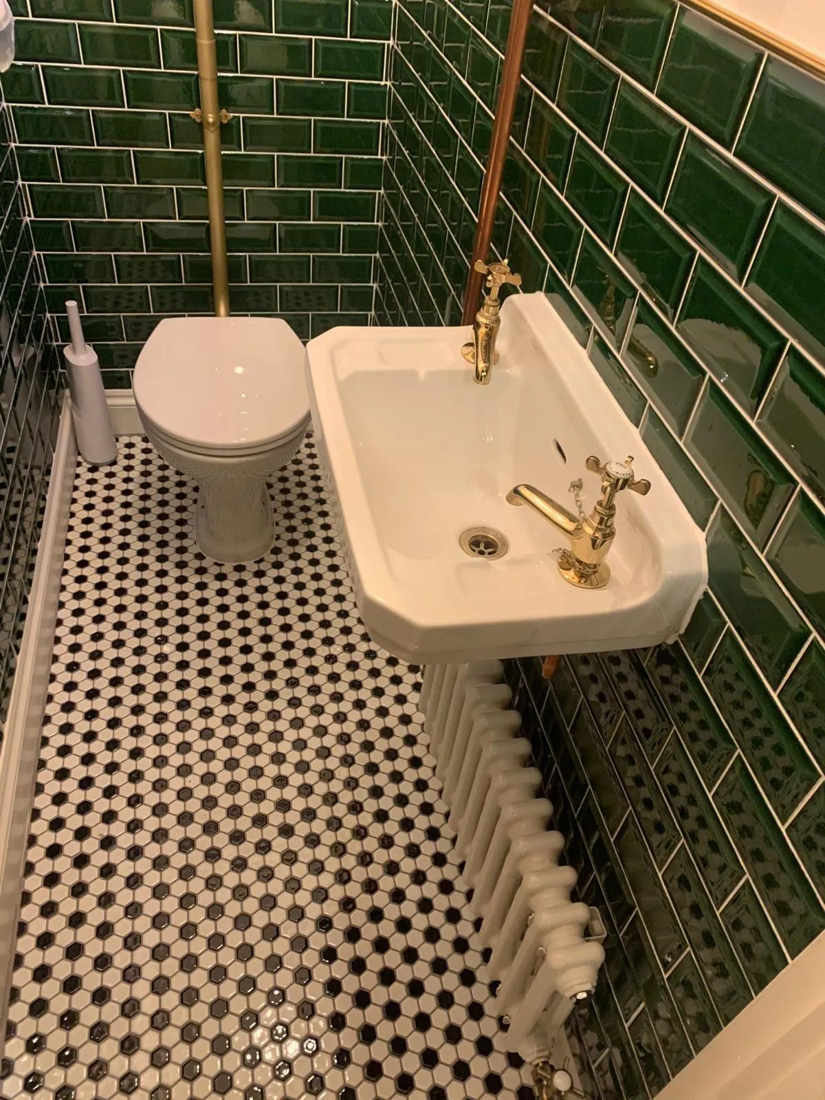A wall-hung basin with a brass tap and a toilet against dark green metro tiling, above a patterned tiled floor
