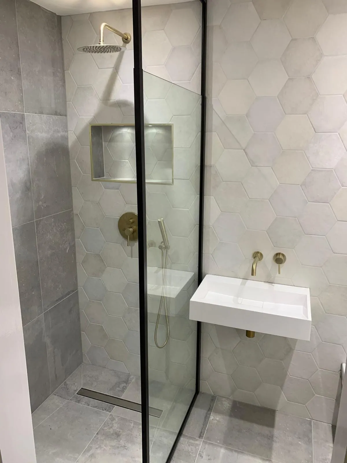 A walk-in shower behind a frameless glass screen, with hexagonal wall tiles and brushed brass fittings