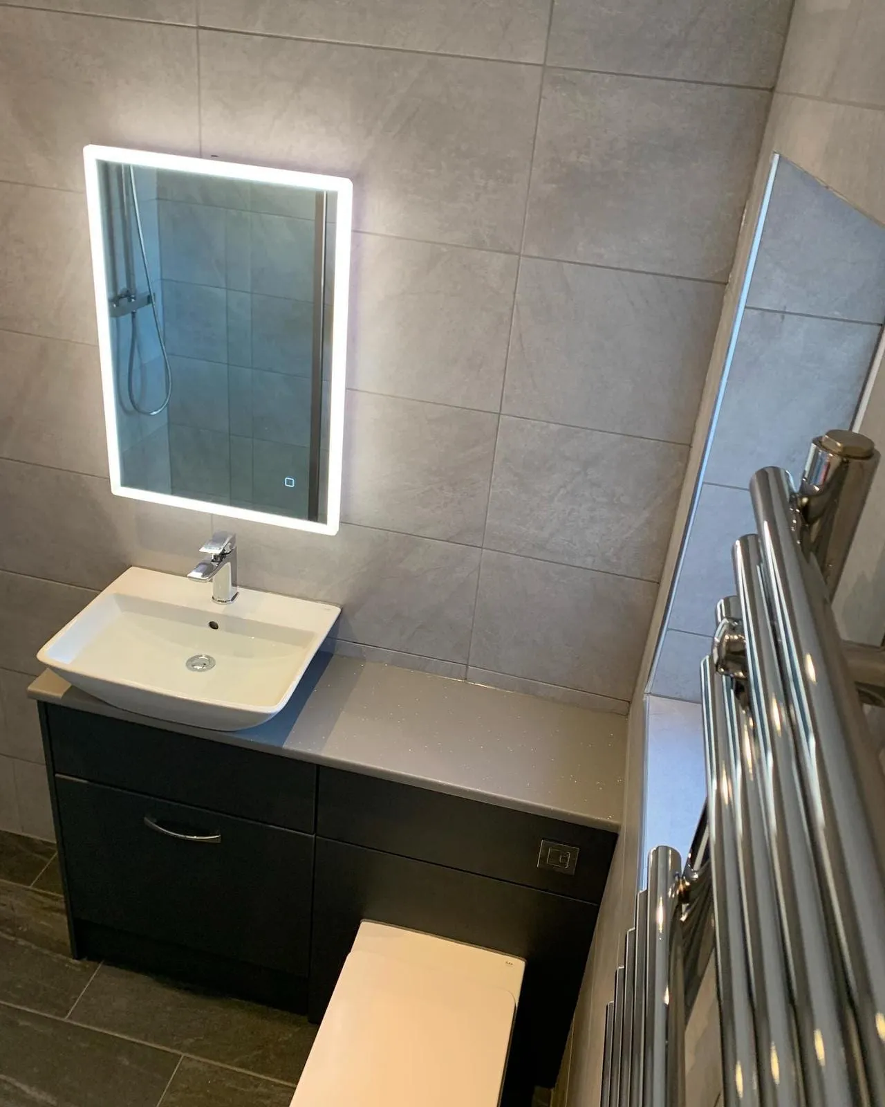 A backlit mirror above a dark vanity unit, alongside a chrome towel rail