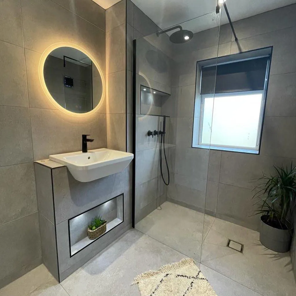 A round backlit mirror above a wall-hung basin unit, next to a tiled walk-in shower and a window