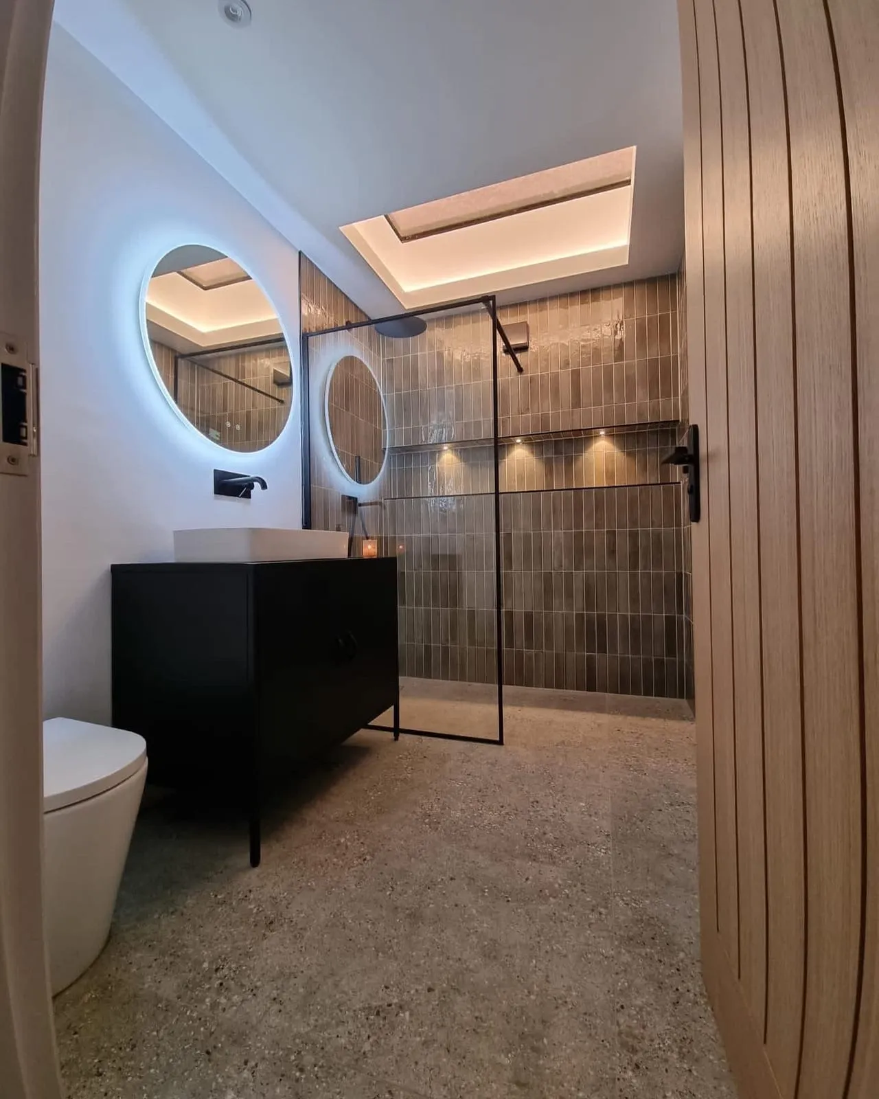 A finished bathroom with twin round backlit mirrors above a black vanity unit, a timber slat wall and a walk-in shower