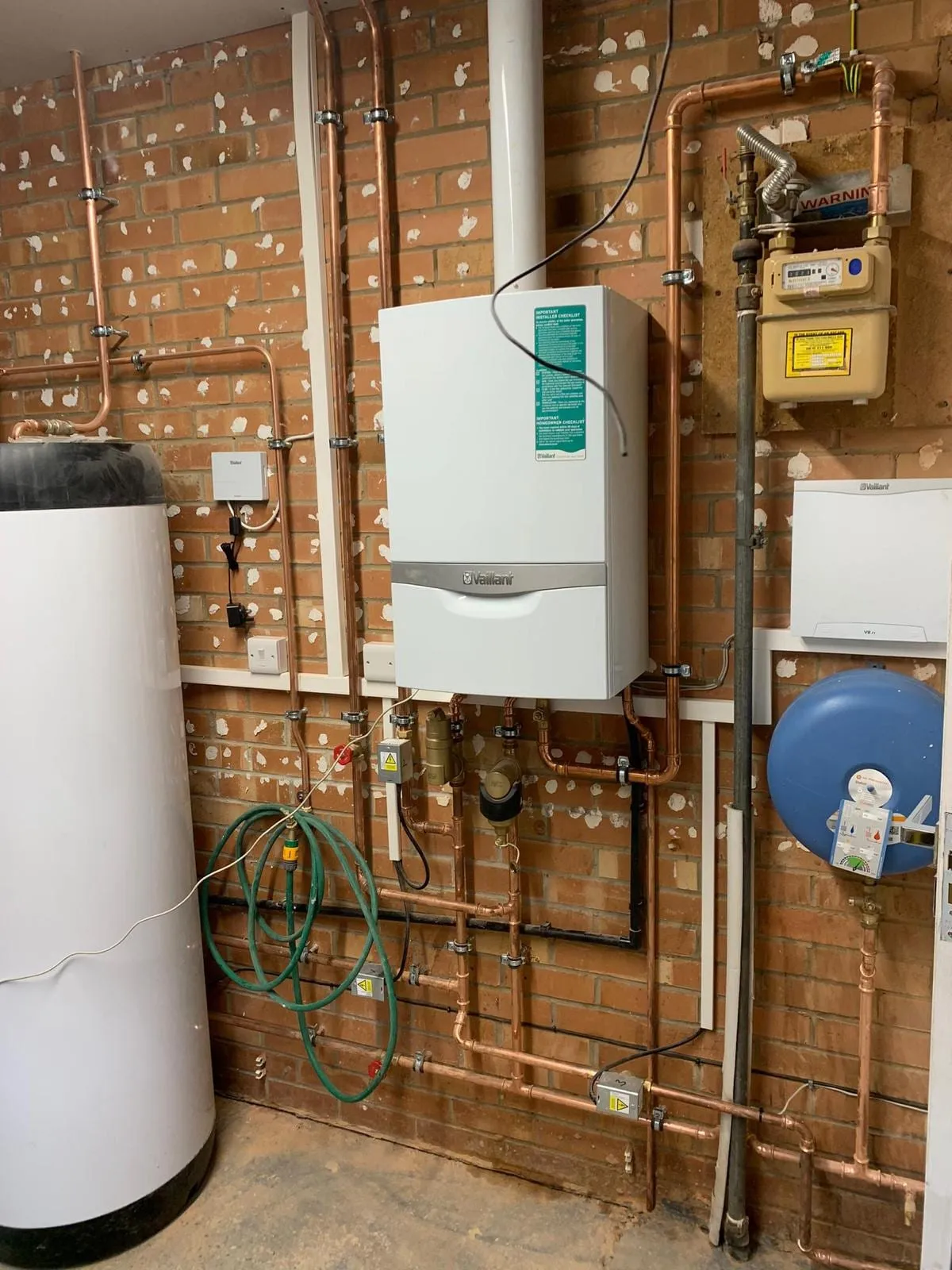 A boiler and hot water cylinder mounted on a brick wall with the copper pipework run between them