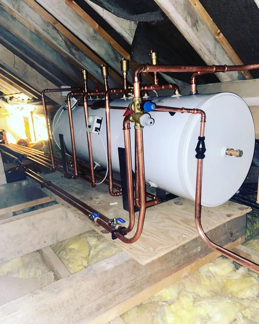 A hot water cylinder installed in a loft with copper pipework run across the joists