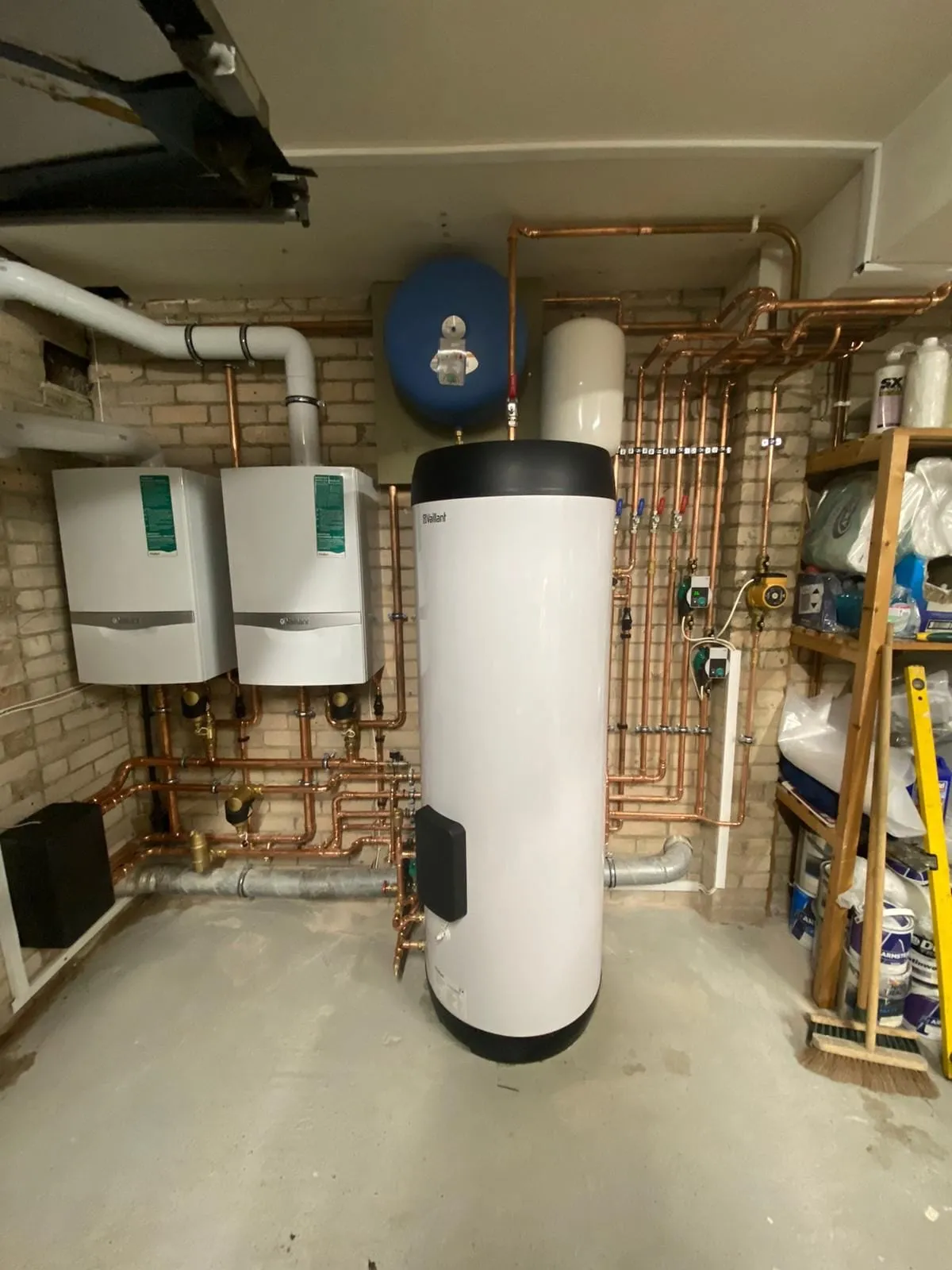 Two boilers and a large hot water cylinder in a plant space, with the pipework and controls laid out on the wall