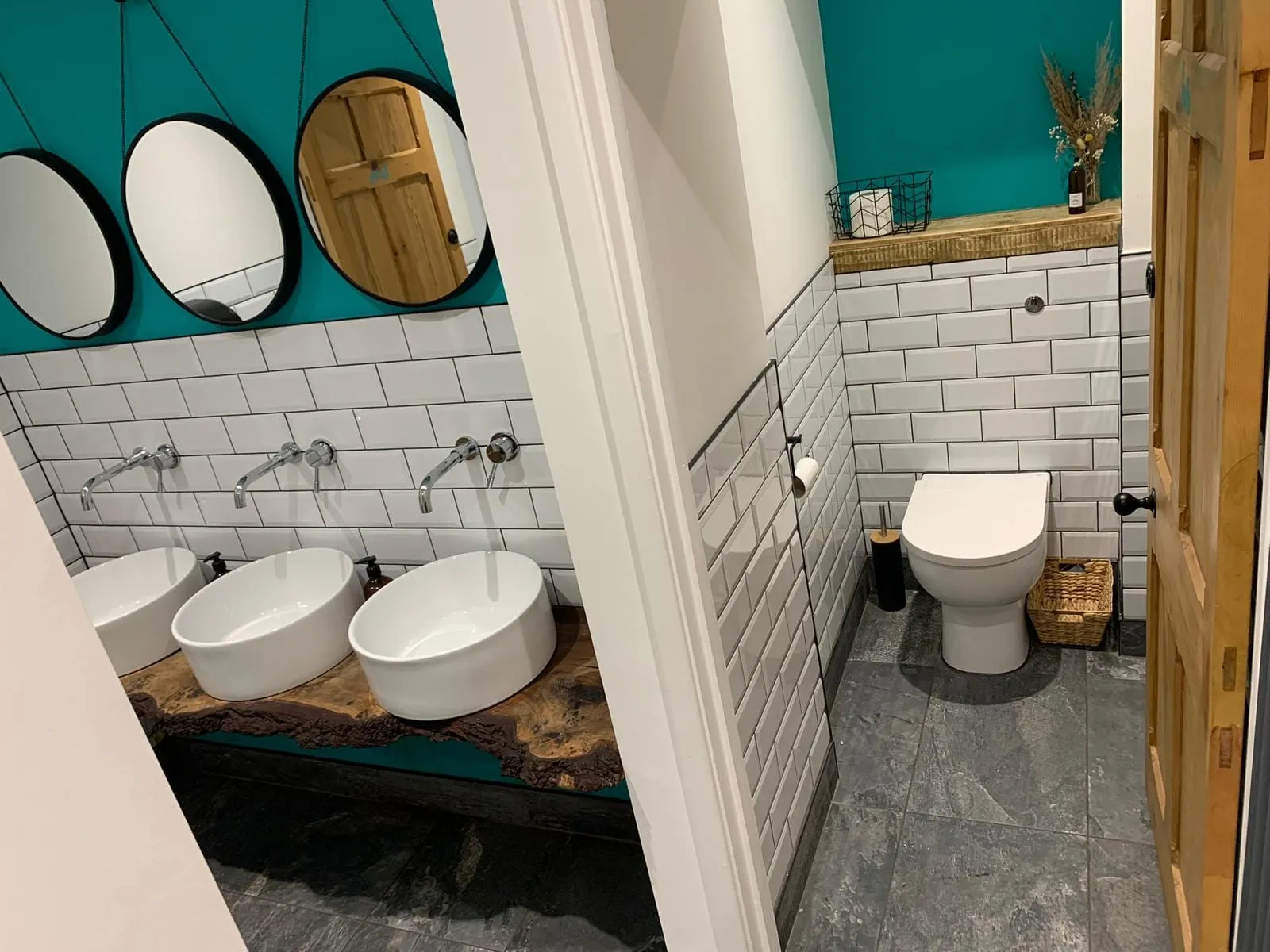 A commercial washroom with counter-top basins on a timber shelf, round mirrors and a toilet cubicle beyond